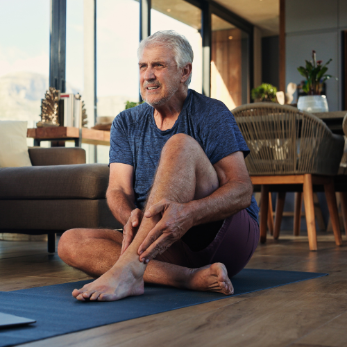 Older man seated on a mat at home holding his ankle and lower leg due to pain and discomfort.
